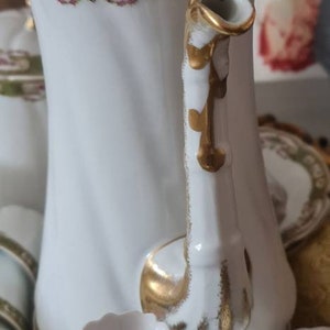 May include: White porcelain tea set with gold accents and floral detailing. The set includes a teapot with a lid, cups, and saucers. The teapot handle and lid finial are gold. The set has a vintage aesthetic.