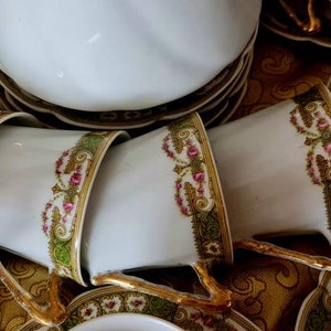 May include: A collection of white porcelain teacups and saucers with gold trim and floral designs. The cups are stacked, showcasing the intricate details of the gold handles and the delicate floral patterns in pink and green.