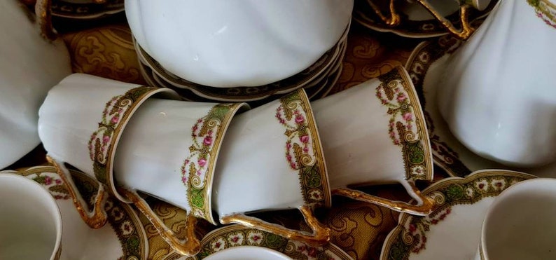 May include: A collection of white porcelain teacups and saucers with gold trim and floral designs. The cups are stacked, showcasing the intricate details of the gold handles and the delicate floral patterns in pink and green.