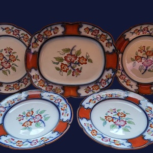 Può includere: Set di cinque piatti in porcellana bianca con bordi arancioni, blu e dorati. Ogni piatto presenta un motivo floreale con fiori rosa e verdi.