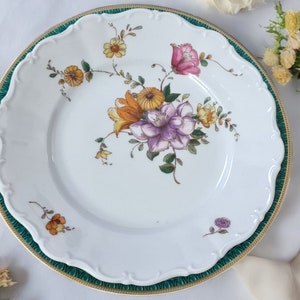 Puede incluir: Un plato de porcelana blanca con un borde verde y dorado. El plato presenta un diseño floral con flores amarillas, rosas y moradas.
