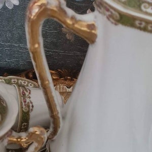 May include: White porcelain tea set with gold accents. The set includes a teapot, cups, and saucers, all adorned with a delicate floral and gold trim design. The teapot handle is gold. The background features a floral painting.