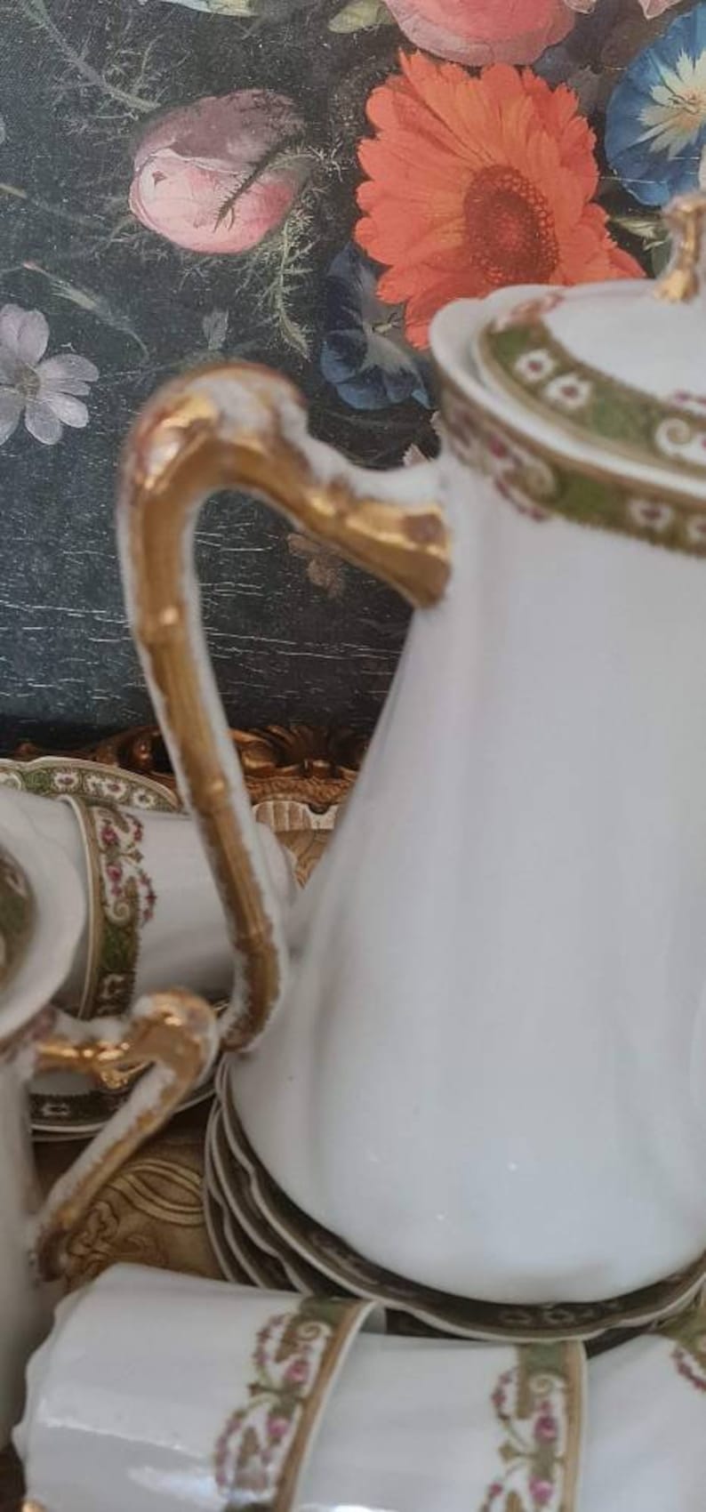 May include: White porcelain tea set with gold accents. The set includes a teapot, cups, and saucers, all adorned with a delicate floral and gold trim design. The teapot handle is gold. The background features a floral painting.