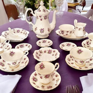 Puede incluir: Un juego de té floral morado con una tetera, una jarra de crema, un azucarero y cuatro tazas y platillos. El juego está sobre un mantel morado.