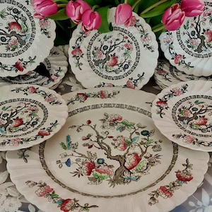 Pode incluir: Um conjunto de pratos de porcelana vintage com um padrão floral com uma árvore e flores cor-de-rosa. Os pratos são brancos com uma borda marrom e estão dispostos em um padrão circular.