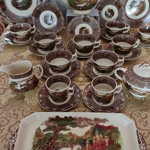 May include: A vintage tea set with a brown and white floral pattern. The set includes a teapot, creamer, sugar bowl, and six teacups and saucers. The teacups and saucers have a brown and white floral pattern with a green and brown landscape scene. The teapot has a brown and white floral pattern with a green and brown landscape scene. The creamer and sugar bowl have a brown and white floral pattern with a green and brown landscape scene.