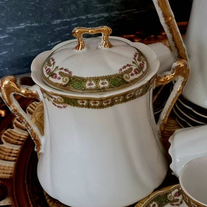 May include: White porcelain sugar bowl with a lid, featuring gold accents on the handles and rim. The lid and rim are decorated with a floral and green design. The sugar bowl is part of a tea set.