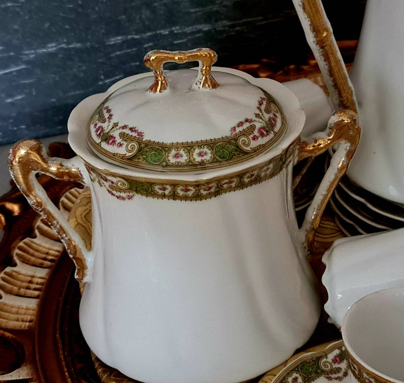 May include: White porcelain sugar bowl with a lid, featuring gold accents on the handles and rim. The lid and rim are decorated with a floral and green design. The sugar bowl is part of a tea set.