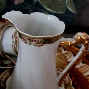 May include: White porcelain creamer with a scalloped rim and gold accents. The handle is gold, and the rim features a decorative band with red and green floral patterns. A matching teacup and saucer are also visible.