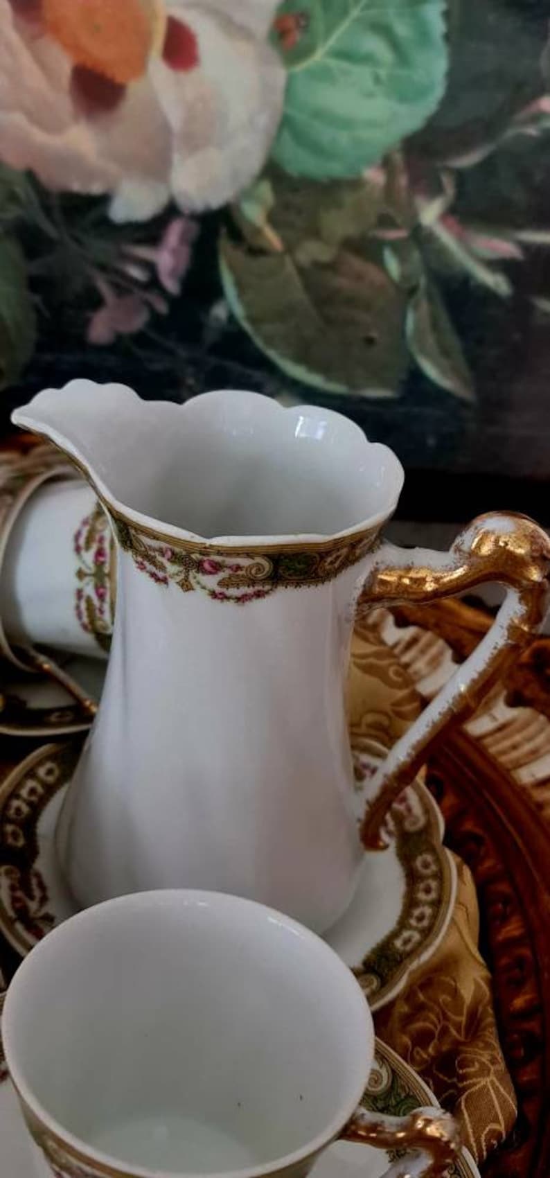 May include: White porcelain creamer with a scalloped rim and gold accents. The handle is gold, and the rim features a decorative band with red and green floral patterns. A matching teacup and saucer are also visible.