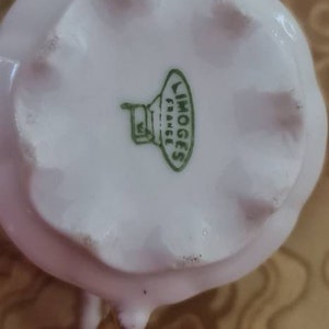 May include: A close-up of a white porcelain creamer with a scalloped edge. The bottom of the creamer has a green stamp that reads "LIMOGES FRANCE". The creamer has a small spout and a handle.