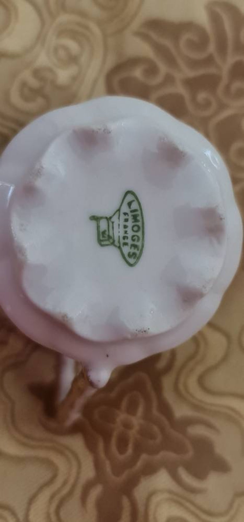 May include: A close-up of a white porcelain creamer with a scalloped edge. The bottom of the creamer has a green stamp that reads "LIMOGES FRANCE". The creamer has a small spout and a handle.