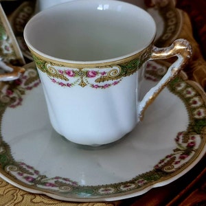 May include: White teacup and saucer set with gold trim and floral accents. The cup has a gold handle and a decorative band of green, gold, and pink floral designs. The saucer has a scalloped edge and matching trim.