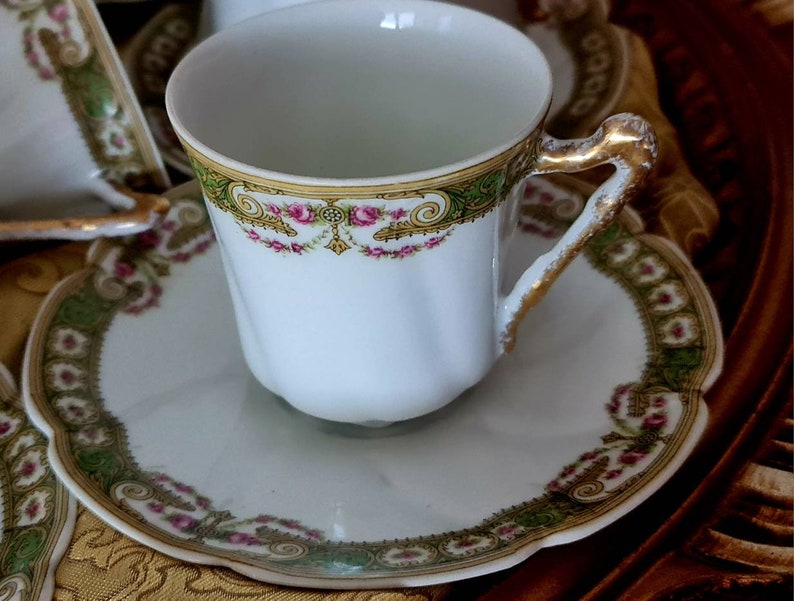 May include: White teacup and saucer set with gold trim and floral accents. The cup has a gold handle and a decorative band of green, gold, and pink floral designs. The saucer has a scalloped edge and matching trim.