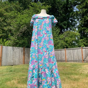 May include: A turquoise maxi dress with a floral pattern. The dress features a ruffled neckline and hem, short sleeves, and a vibrant print of pink, purple, and yellow flowers and leaves. The dress is displayed on a mannequin outdoors.