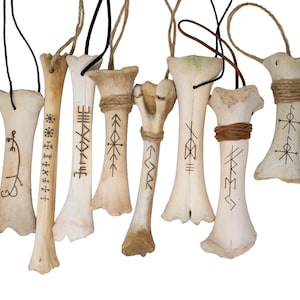 May include: A collection of eight off-white bone pendants, each featuring unique black ink symbols or runes. Several bones are wrapped with thin brown or black twine, and each piece includes a cord for hanging, creating a set of decorative ornaments.