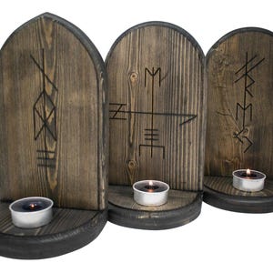 Puede incluir: Tres candelabros de madera con acabado marrón oscuro. Cada soporte presenta un símbolo de runa tallado y una pequeña vela de té encendida. Los soportes tienen un aspecto rústico y envejecido.