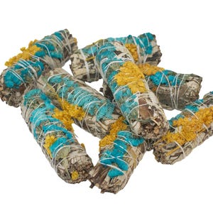 Pode incluir: Um feixe de varinhas de sálvia branca envoltas em barbante azul e amarelo. As varinhas são amarradas juntas com um barbante branco.