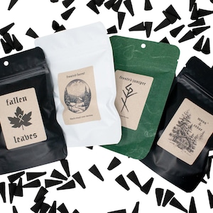 Puede incluir: Cuatro bolsas de incienso en negro, blanco y verde, cada una con una etiqueta. Las etiquetas presentan una hoja de arce, una escena de bosque, una runa y una ilustración de pino. Conos de incienso negro dispersos.