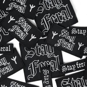 Op de afbeelding: Zwarte geborduurde patches met de tekst "Stay Feral" in een gotisch lettertype. Sommige patches bevatten een klein symbool. De patches zijn in verschillende maten en vormen, verspreid op een witte ondergrond.