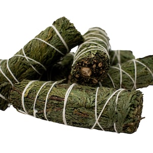Puede incluir: Un conjunto de manojos de salvia verde, atados con hilo blanco. Los manojos están hechos de hojas y tallos secos, y están dispuestos en una pila sobre un fondo blanco. La salvia tiene un aspecto natural y terroso.