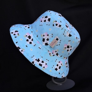 May include: A light blue bucket hat with a black and white cow print design. The hat has a small label that says "Hand-Made by PoppyBliss".