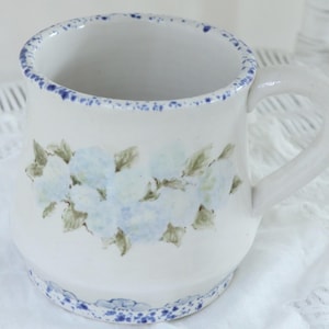 May include: White ceramic mug with a blue floral design and a blue rim. The mug features a curved handle and is decorated with light blue flowers and green leaves. The base and rim have a speckled blue trim. The mug is sitting on a white surface.