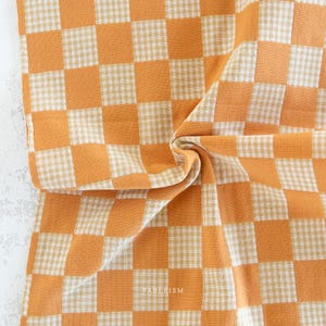 May include: A textile fabric featuring a large-scale checkerboard pattern in shades of orange and white. Each large square is composed of a smaller, fine gingham check. The word FABLEISM is printed in small white letters near the bottom center.