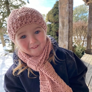 May include: A young child wearing a pink crocheted hat and matching scarf. The hat and scarf are both made with a textured stitch. The child is wearing a dark blue jacket.