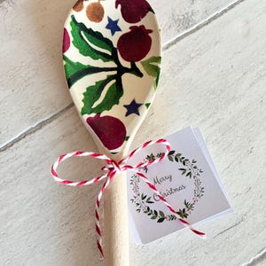 Puede incluir: Una cuchara de madera con un patr&oacute;n festivo de bayas rojas, hojas verdes y estrellas azules en el cuenco. El mango est&aacute; atado con cordel de panadero rojo y blanco, que sujeta una peque&ntilde;a etiqueta cuadrada con una corona y Merry Christmas.