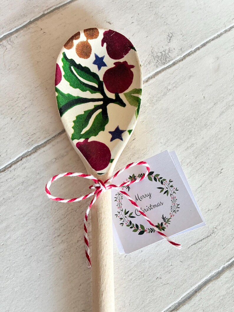 Puede incluir: Una cuchara de madera con un patr&oacute;n festivo de bayas rojas, hojas verdes y estrellas azules en el cuenco. El mango est&aacute; atado con cordel de panadero rojo y blanco, que sujeta una peque&ntilde;a etiqueta cuadrada con una corona y Merry Christmas.