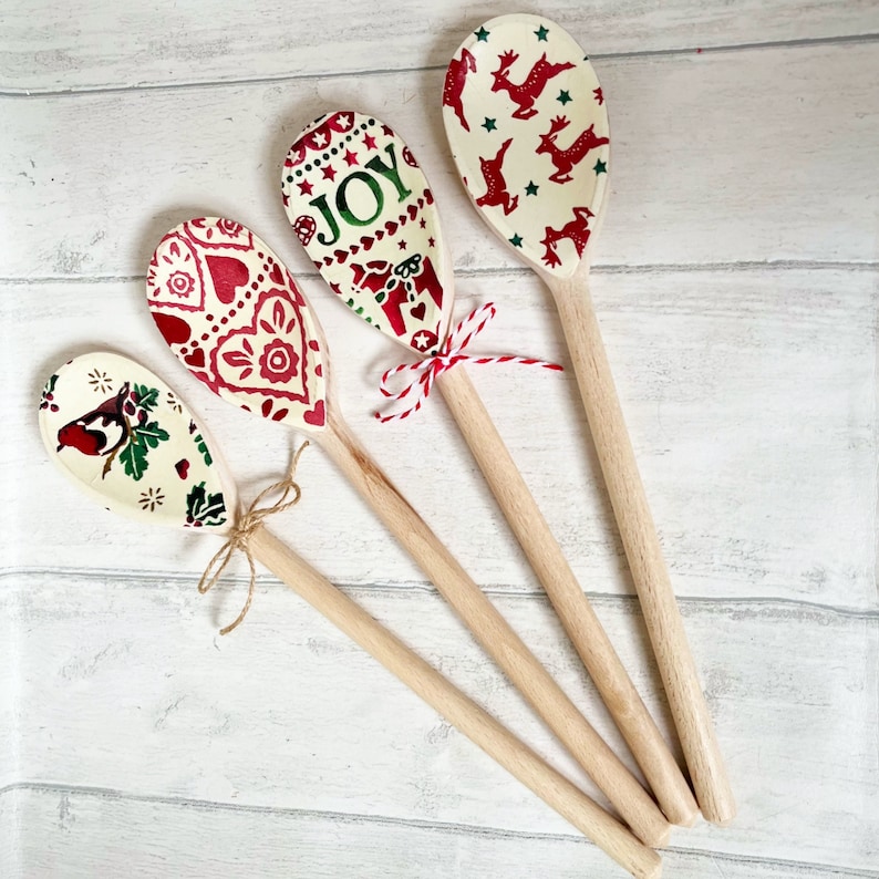 Puede incluir: Cuatro cucharas de madera con mangos de madera clara y cuencos de color crema con dise&ntilde;os festivos. Una cuchara muestra un petirrojo, otra corazones rojos, una incluye la palabra JOY con un reno y la &uacute;ltima muestra renos rojos.
