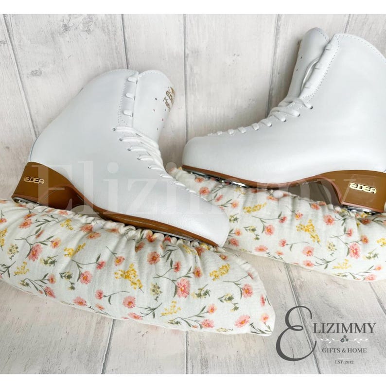 May include: A pair of white figure skates with brown leather soles and gold accents. The skates are on a pair of white floral skate guards.