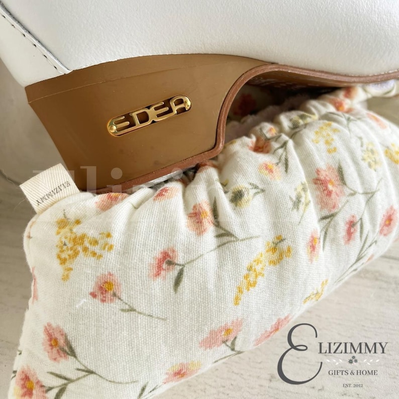May include: White figure skates with a brown sole and a gold EDEA logo. The skates are covered with a white fabric skate protector with a floral pattern in pink and yellow.