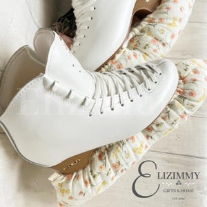 May include: White figure skates with a white lace-up design and a gold logo on the sole. The skates are on top of a pair of white and yellow floral skate guards.