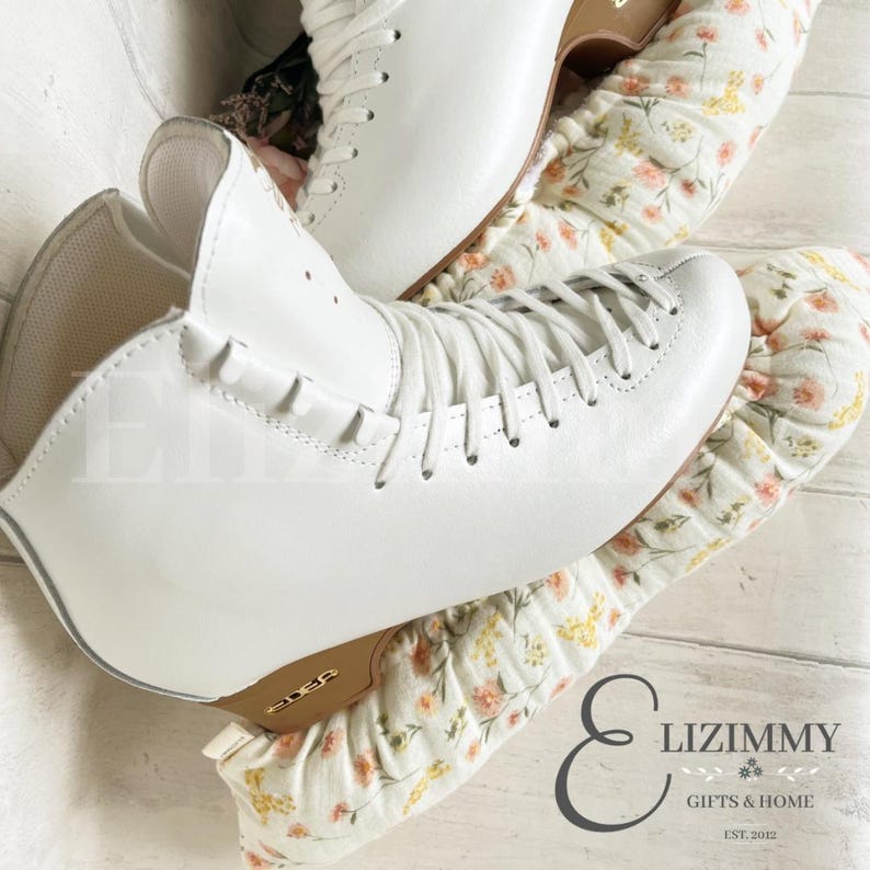 May include: White figure skates with a white lace-up design and a gold logo on the sole. The skates are on top of a pair of white and yellow floral skate guards.