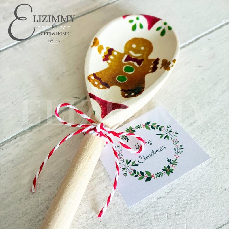 Puede incluir: Una cuchara de madera clara con una ilustraci&oacute;n de un hombre de jengibre marr&oacute;n en el cuenco. El mango est&aacute; atado con un cordel de panadero rojo y blanco sujeto a una etiqueta cuadrada blanca con una corona verde y el texto Merry Christmas.