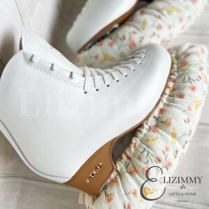 May include: White figure skates with a brown wooden base and gold lettering that reads "Edea". The skates are on top of a white fabric with a floral pattern and pink accents. The fabric has a ruffled edge and the text "Elizimmy Gifts & Home Est. 2012" is printed on the fabric.