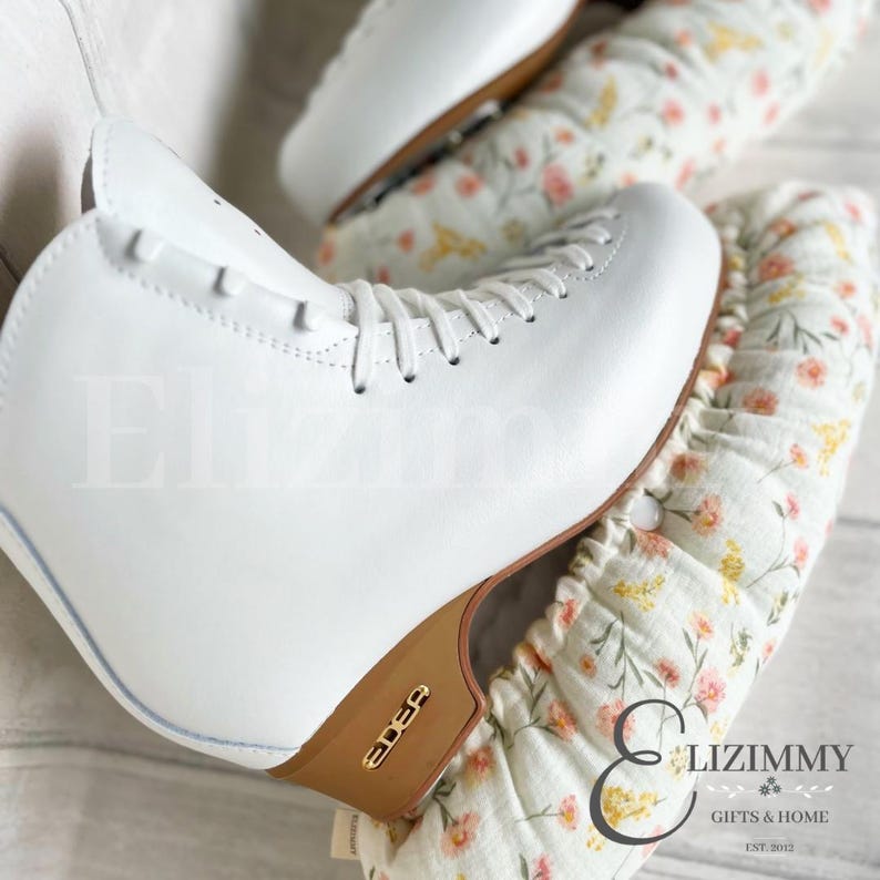 May include: White figure skates with a brown wooden base and gold lettering that reads "Edea". The skates are on top of a white fabric with a floral pattern and pink accents. The fabric has a ruffled edge and the text "Elizimmy Gifts & Home Est. 2012" is printed on the fabric.