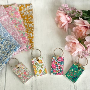 May include: Four floral patterned keychains with silver key rings. The keychains are rectangular and feature different floral patterns in shades of pink, blue, green, and yellow.