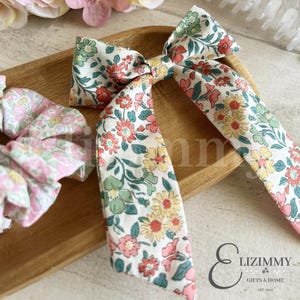 May include: A fabric hair bow and a matching scrunchie, both featuring a white base with a multi-coloured floral pattern in shades of red, yellow, pink, and green. The bow has long, trailing tails, and the scrunchie has a gathered, ruffled texture.