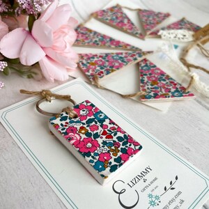 May include: A white ceramic keychain with a floral pattern in pink, blue, and green. The keychain is attached to a silver key ring and a brown twine loop. The keychain is displayed on a white card with the text "Lizimmy Gifts & Home Est. 2012" printed on it.