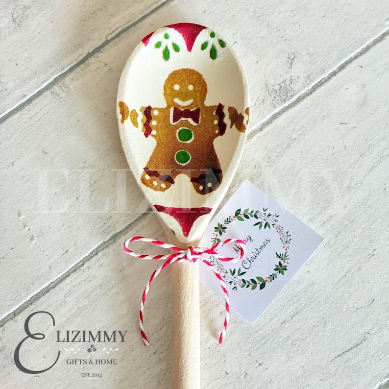 Puede incluir: Una cuchara de madera con un hombre de jengibre pintado a mano en color marr&oacute;n, botones verdes y pajarita roja. La cuchara est&aacute; atada con cordel rojo y blanco e incluye una etiqueta cuadrada con una corona floral y el texto Merry Christmas.