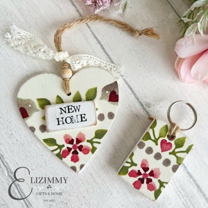 Puede incluir: Un conjunto de dos artículos de decoración para el hogar de madera. Un adorno en forma de corazón con un patrón floral y las palabras "NEW HOME" en una pequeña etiqueta. Un llavero rectangular con un patrón floral similar. Ambos artículos están pintados en tonos de blanco, verde, rosa y gris.