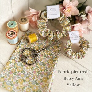 May include: A close-up of a yellow and white floral fabric with a pair of scissors, spools of thread, and two scrunchies made from the same fabric. The fabric is labeled "Betsy Ann Yellow".
