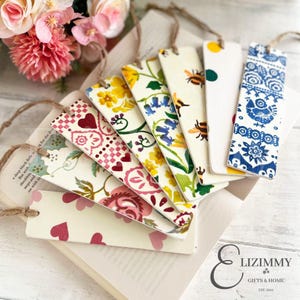 Puede incluir: Un conjunto de marcapáginas decorativos con diversos diseños florales, de corazones e insectos. Cada marcapáginas tiene un diseño único y está sujeto a una cuerda de hilo natural. Los marcapáginas están colocados sobre un libro abierto.