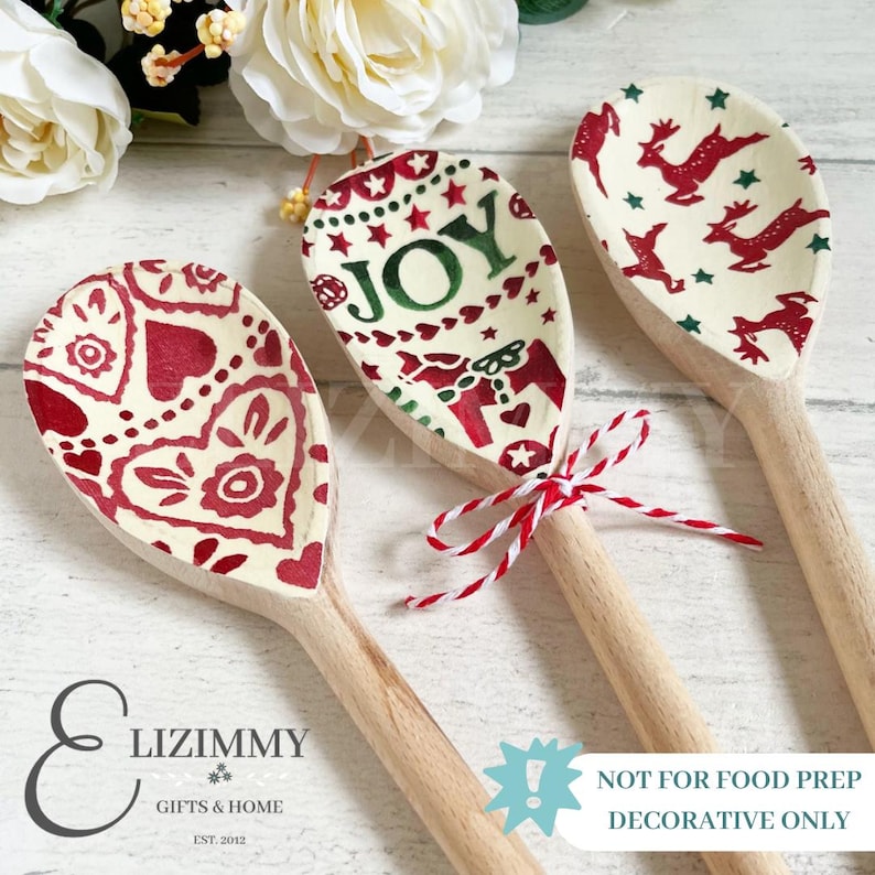 Puede incluir: Tres cucharas de madera con patrones decorativos. La cuchara de la izquierda presenta corazones rojos, la central muestra la palabra JOY con estrellas y un lazo rojo, y la de la derecha tiene renos rojos y estrellas verdes.