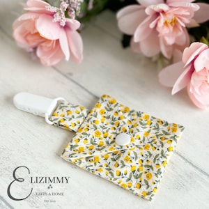 Puede incluir: Un clip blanco con una bolsa de tela floral amarilla y blanca adjunta. La bolsa tiene un cierre de botón blanco. El texto "Lizimmy Gifts & Home Est. 2012" es visible en la esquina inferior izquierda de la imagen.