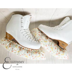 May include: A pair of white figure skates with brown blades are on top of white fabric skate guards with a floral pattern. The guards have a gathered ruffle around the edges.