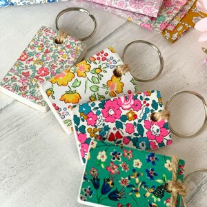 May include: Four keychains with different floral patterns. Each keychain has a silver ring and a brown twine loop. The patterns include a white background with pink, yellow, and blue flowers, a white background with pink and blue flowers, a white background with yellow and blue flowers, and a green background with blue, pink, and white flowers.